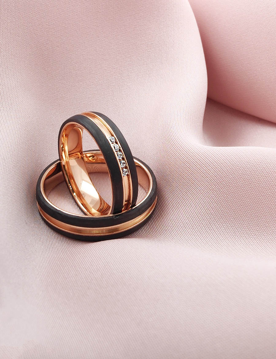 dos anillos originales hechos de oro rosa y carbono. En la parte exterior hay dos bandas de carbono y en medio el oro. Son anillos de bodas bonitos sobre una tela de color rosa
